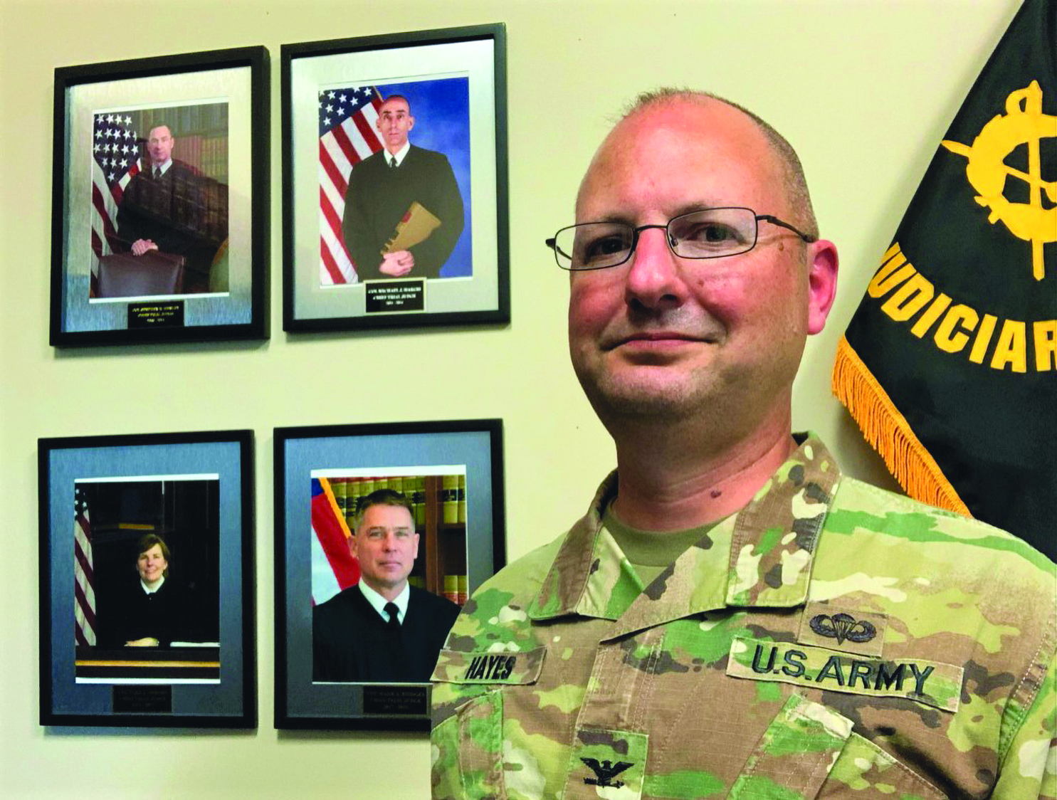 Chief Trial Judge Colonel Timothy Hayes stands next to photos of the previous four Chief Trial Judges:
        Colonel Stephen Henley (upper left), Colonel Michael Hargis (upper right), Colonel Tara Osborn (lower left),
        and Colonel Mark Bridges (lower right). (Photo courtesy of author)